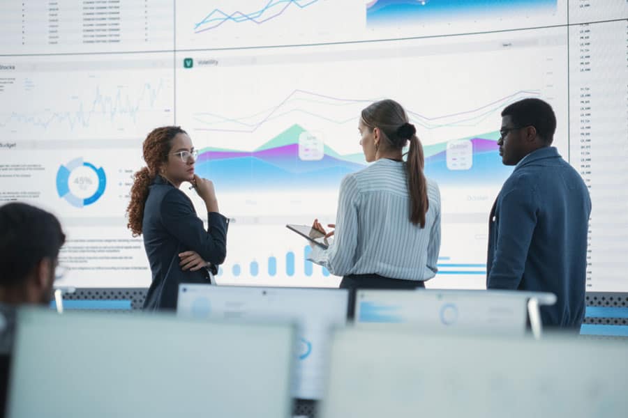 Group of people talking in front of wall of screens with business charts showing