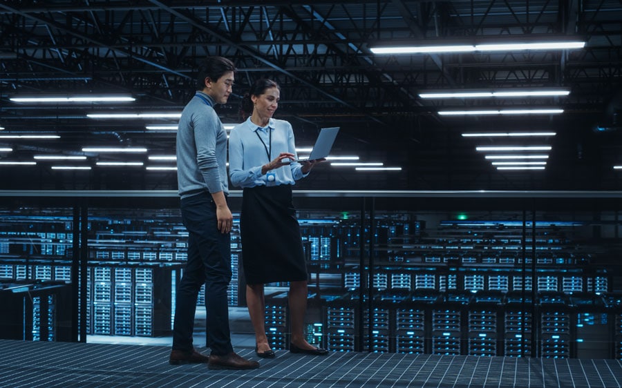 two workers standing on raised platform in data center looking at laptop